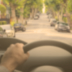 Image from drivers perspective in a car, showing top of wheel in the foreground and road ahead, including signs, blurred and colour dulled down to show vision with cataracts